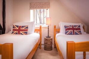 A bedroom with two single beds, each with a pillow featuring the Union Jack design. There is a wooden headboard, a small wooden stool, and a window with a curtain.