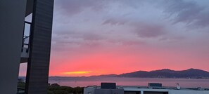 Une vue sur le coucher de soleil, prise depuis un bâtiment donnant sur une étendue d’eau, avec des montagnes lointaines en arrière-plan.