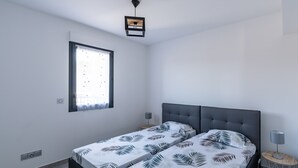 Ein modernes Schlafzimmer mit zwei Betten, einem Fenster mit Jalousien, einer Deckenleuchte und einem kleinen Nachttisch mit Lampe.