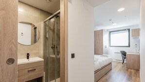 Quarto moderno com chuveiro de vidro, toucador de madeira e cama com escrivaninha.