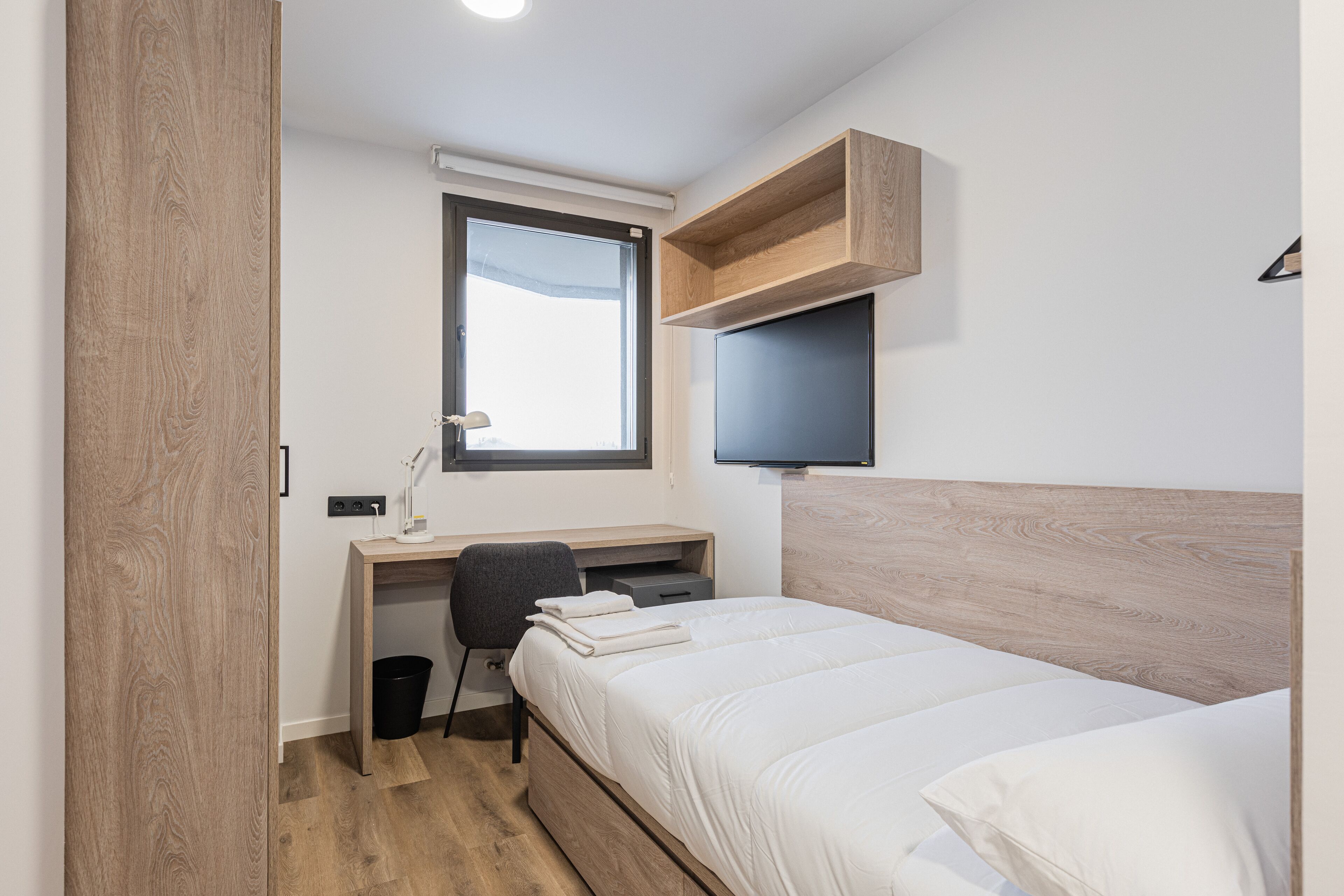 A bedroom with a bed, a desk, a chair, a TV, and a window.