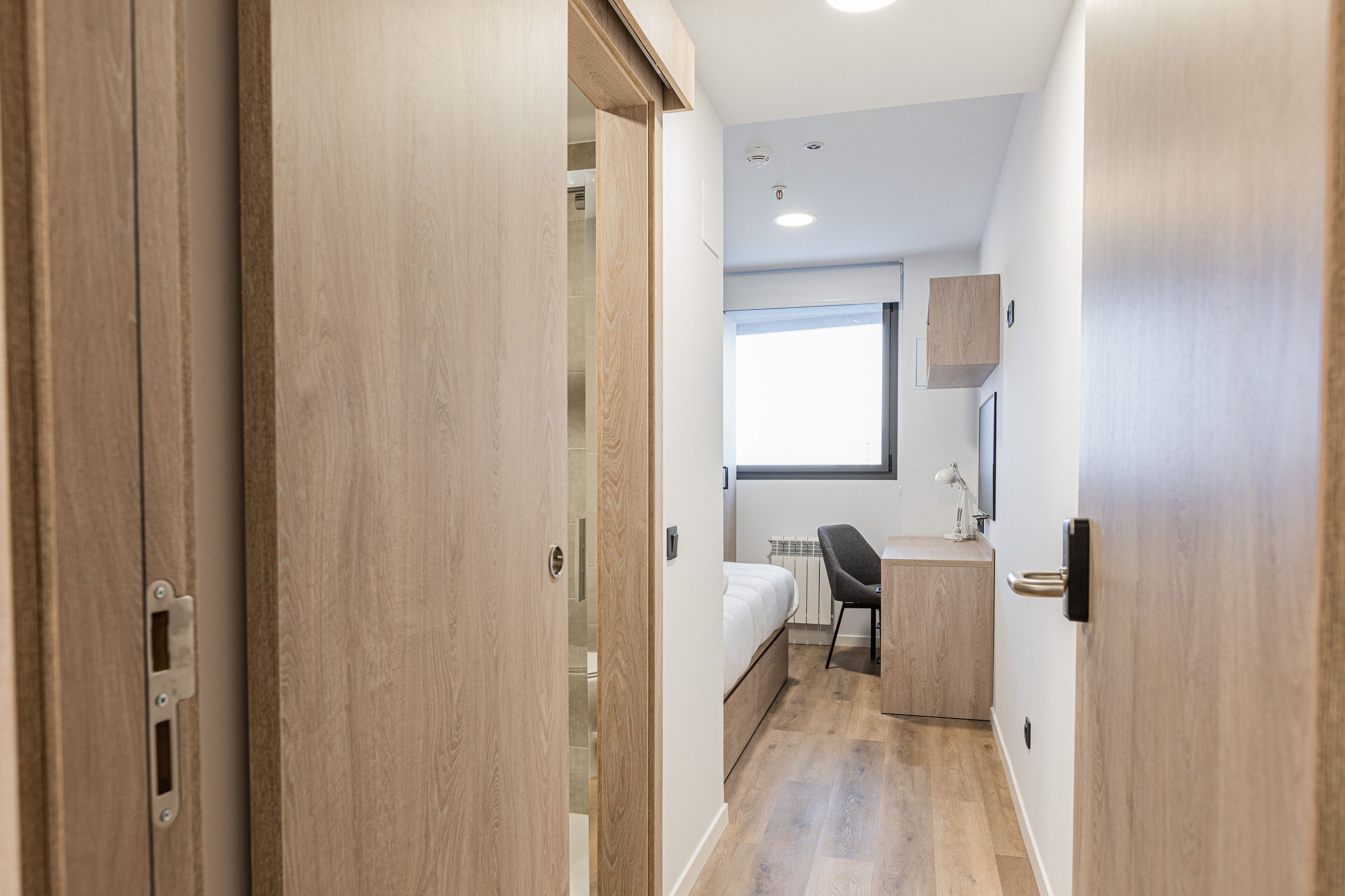 Un pasillo estrecho con puertas de madera, una cama, un escritorio y una silla en una habitación pequeña.