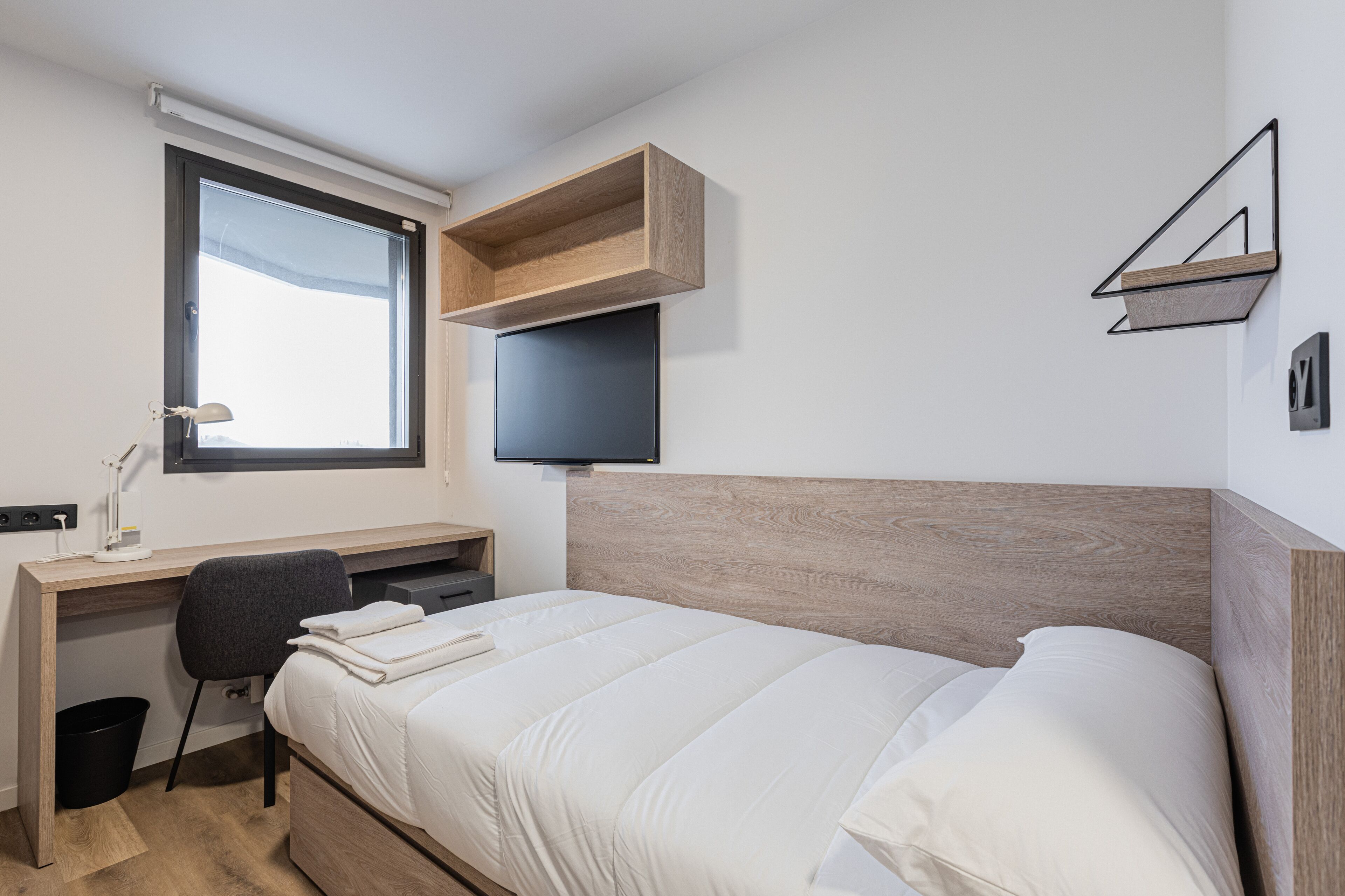 A modern bedroom with a bed, a desk, a chair, and a TV mounted on the wall.