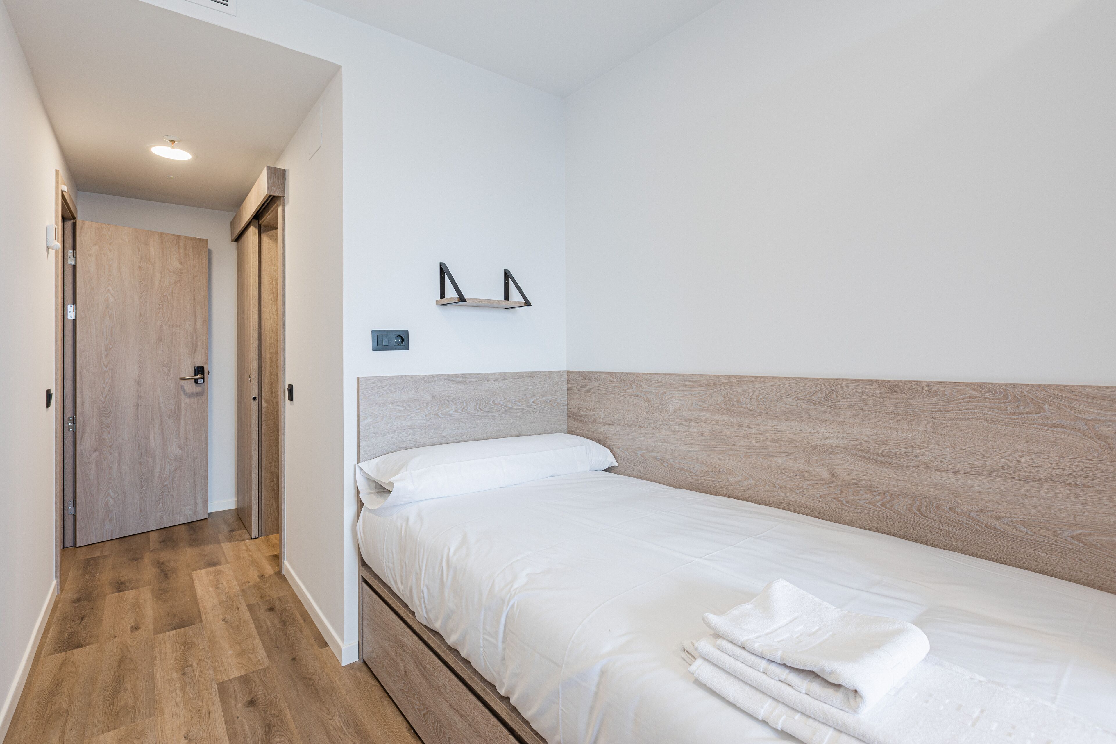 Un dormitori amb un capçal de llit de fusta, un llit amb roba de llit blanca, una petita estant amb un quadre, i una porta que dona a una altra habitació.