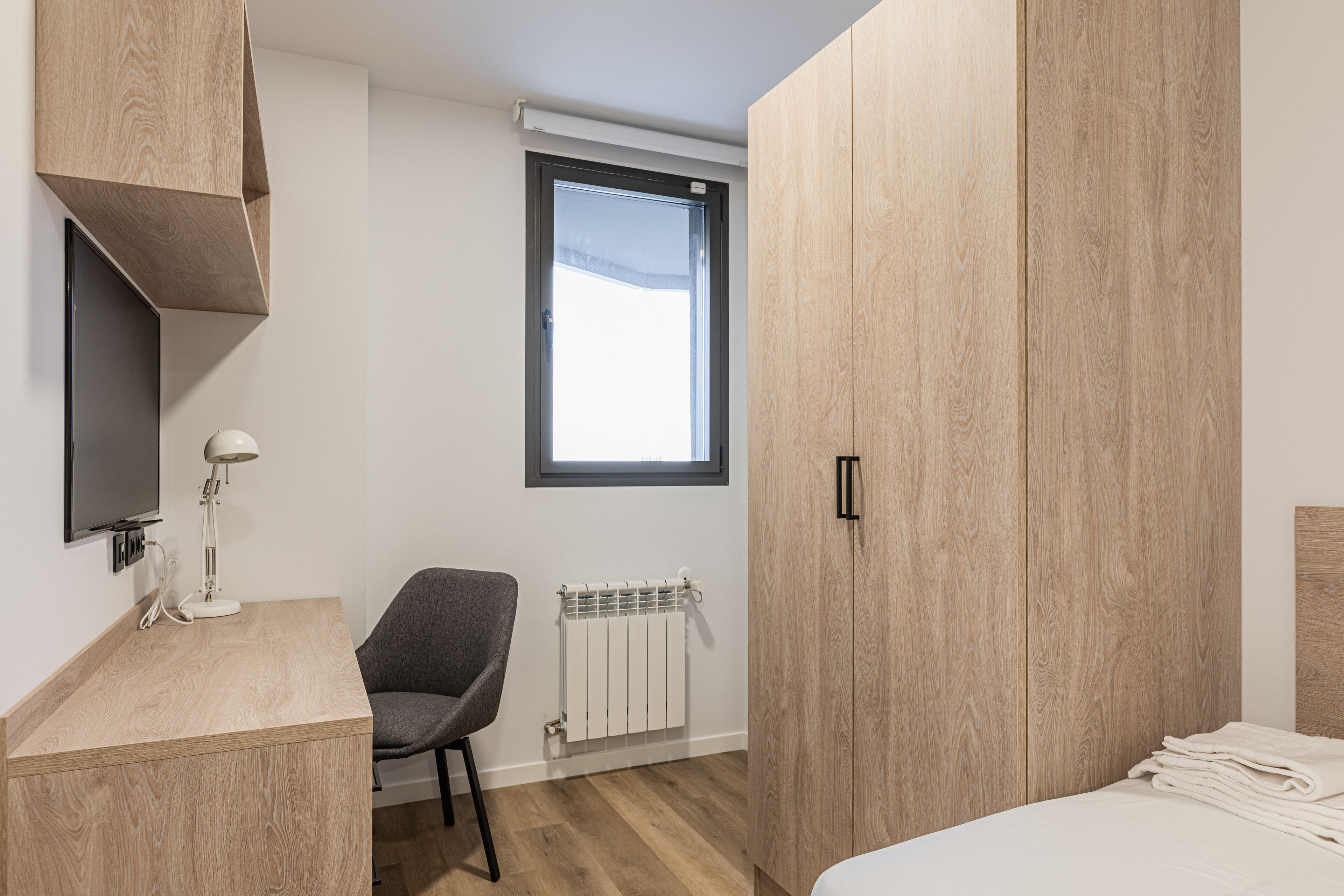 Un dormitorio moderno con un armario de madera, un escritorio con lámpara, una silla, una cama con toallas ordenadas, un radiador y una ventana con vistas.