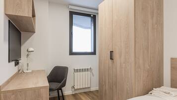 Un dormitorio moderno con un armario de madera, un escritorio con lámpara, una silla, una cama con toallas ordenadas, un radiador y una ventana con vistas.
