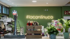 Interior de café com um letreiro de neon com a inscrição "#lacantinucca", máquinas de expresso e plantas em vasos.
