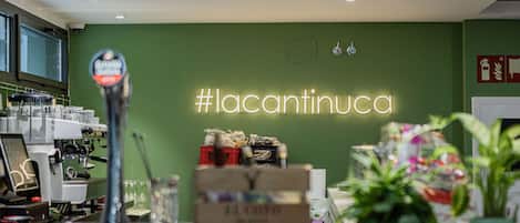 Interior de una cafetería con un letrero de neón que dice '#lacantinucca', máquinas de espresso y plantas en macetas.