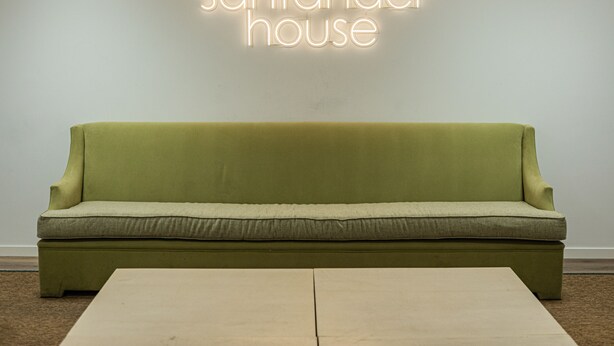 Um sofá verde com uma almofada de cor clara em frente a uma parede com uma placa de neon que diz "santander house".