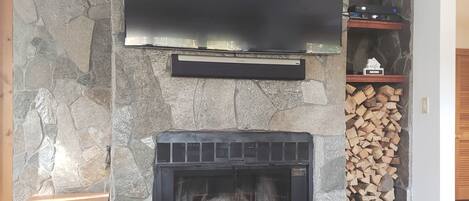 A stone fireplace with a TV mounted above it, a wooden bench, and a stack of firewood to the right.