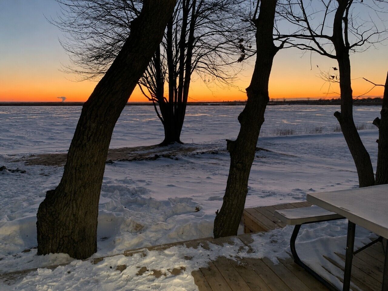 A snowy outdoor area with a wooden deck and a table, surrounded by trees.