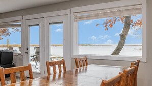 Een eethoek met een grote tafel en stoelen, met uitzicht op een strand en boom door de ramen.