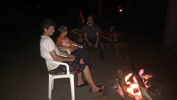 Trois personnes sont assises sur des chaises en plastique, près d’un feu de camp, au milieu de la nuit.