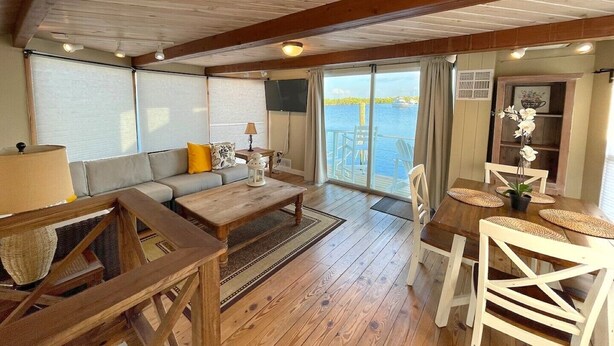 A cozy cabin interior with a wooden floor, a sofa, a dining table with chairs, and a large window overlooking a body of water. - Aqua Lodge Houseboat "Captain's Quarters" (Tavernier)