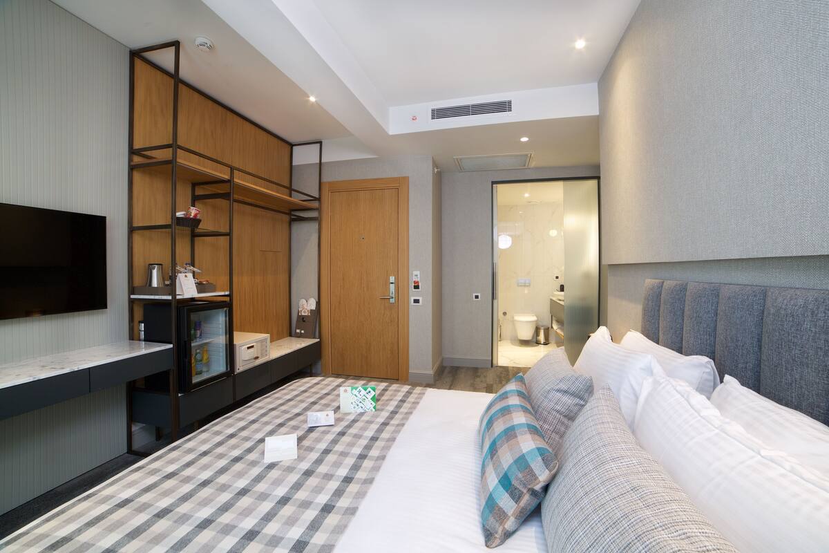 A modern hotel room with a large bed, a flat-screen TV, a wooden wardrobe, and a bathroom visible through an open door.