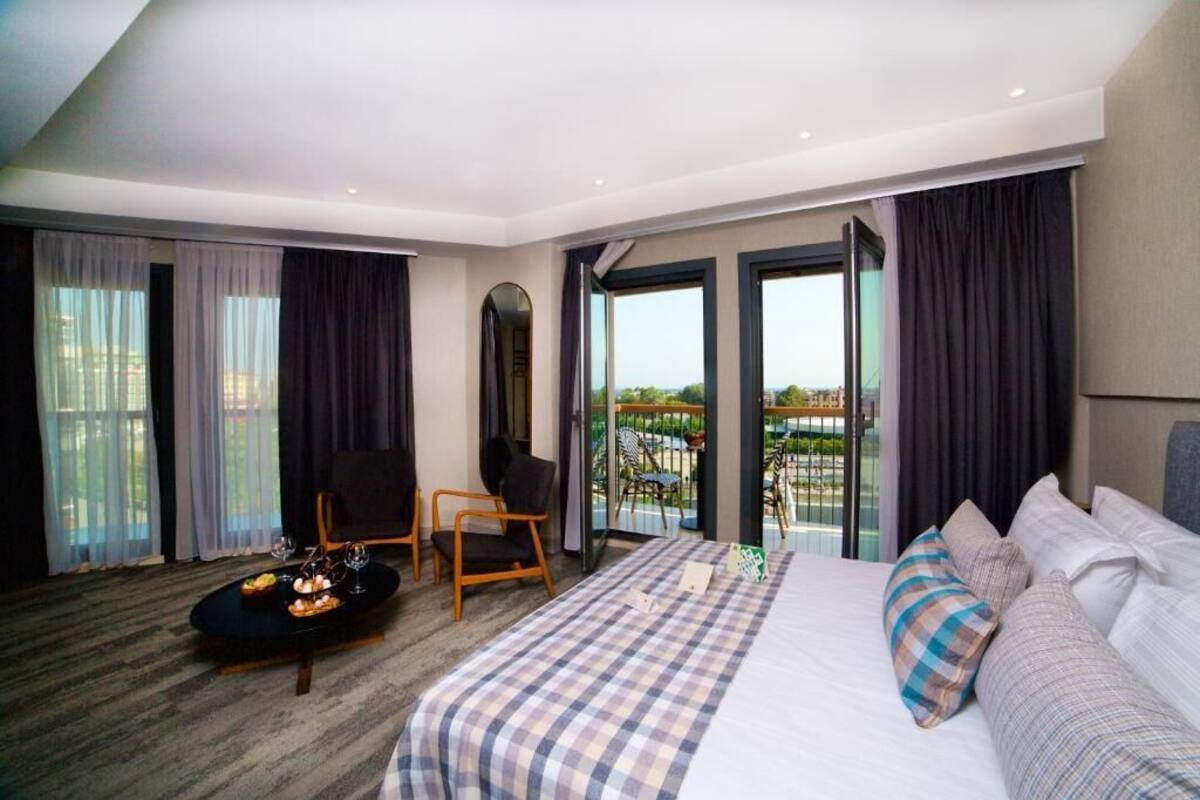 A modern hotel room with a large bed, a small round table with a drink, a chair, and a balcony with a view.