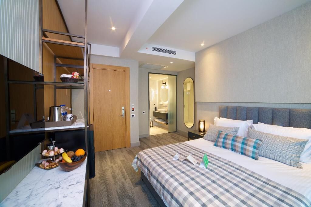 Quarto de hotel moderno com uma cama grande, um guarda-roupa de madeira, uma pequena cozinha e um banheiro visível através de uma porta aberta.