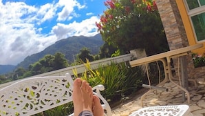 Une personne se détend sur une chaise en métal blanche, avec vue sur une maison, de la verdure et des montagnes en arrière-plan.