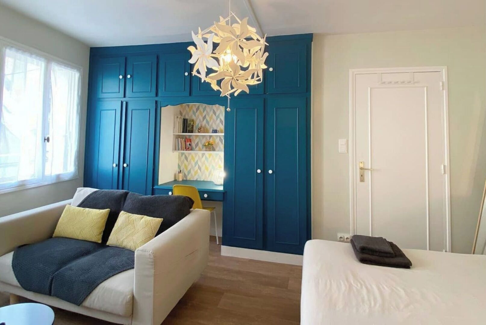 A room with a blue wardrobe, a white sofa with cushions, a small table, and a bed.