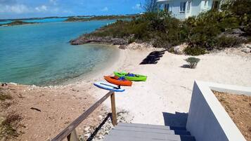A beach view with a staircase leading to a house, kayaks on the sand, and clear blue water.