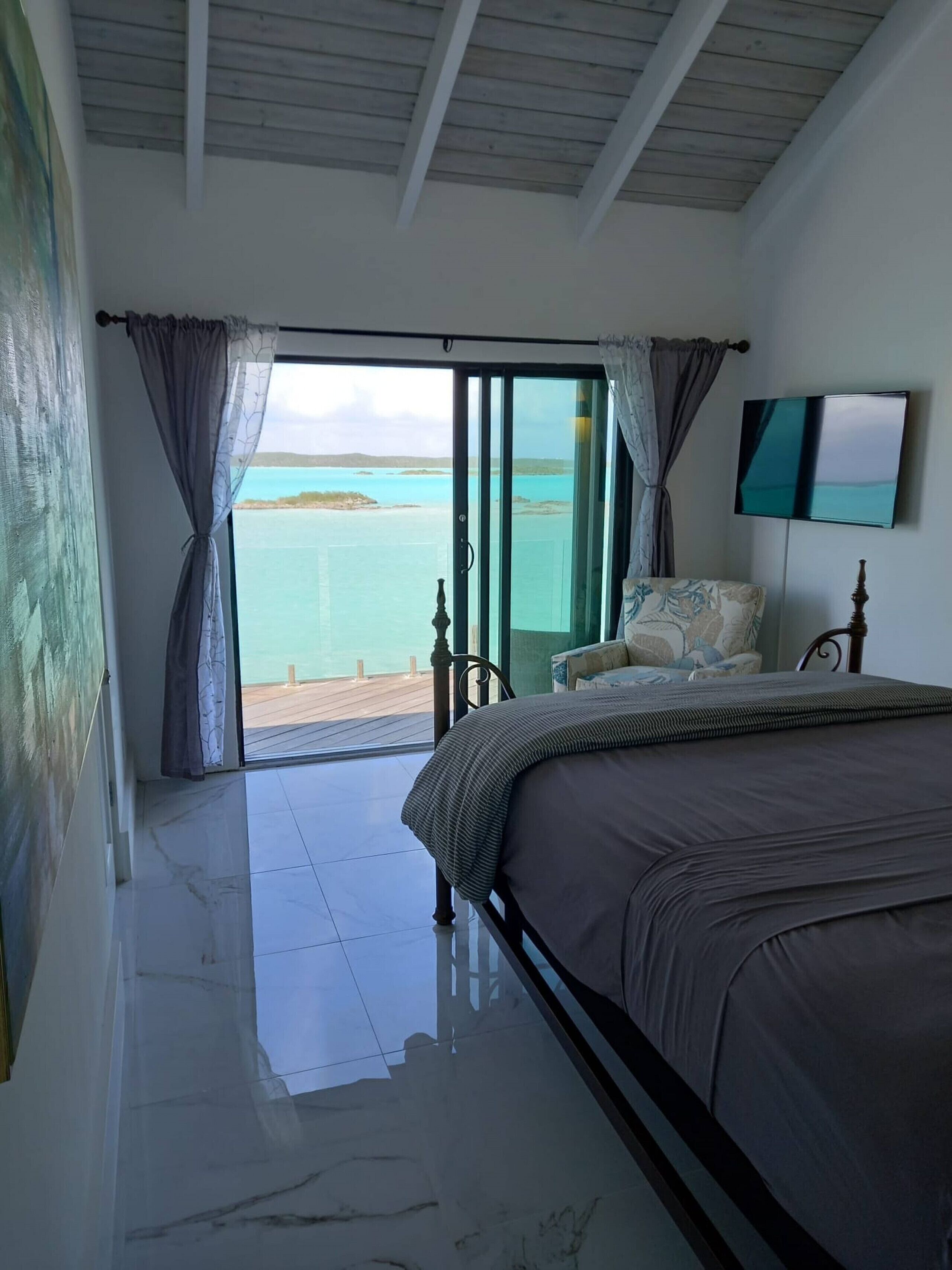 A bedroom with a bed, a chair, a TV, and a view of the sea.