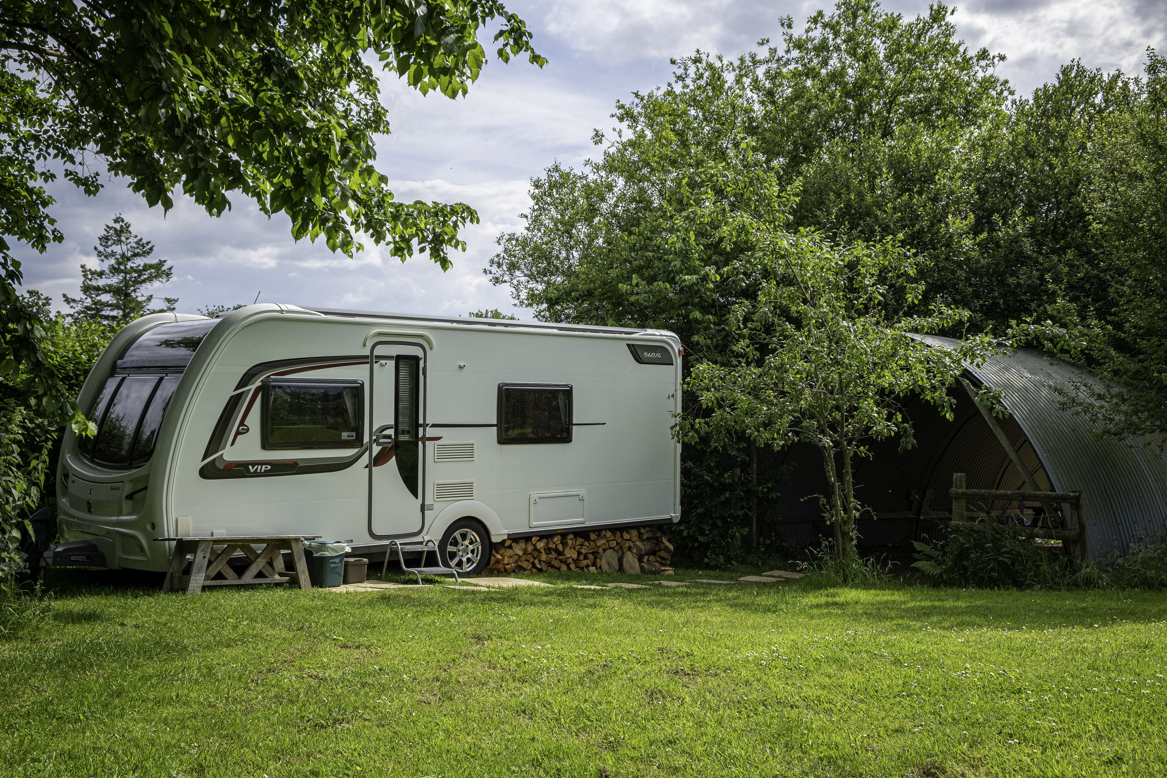 A white RV parked in a grassy area with a covered structure nearby.