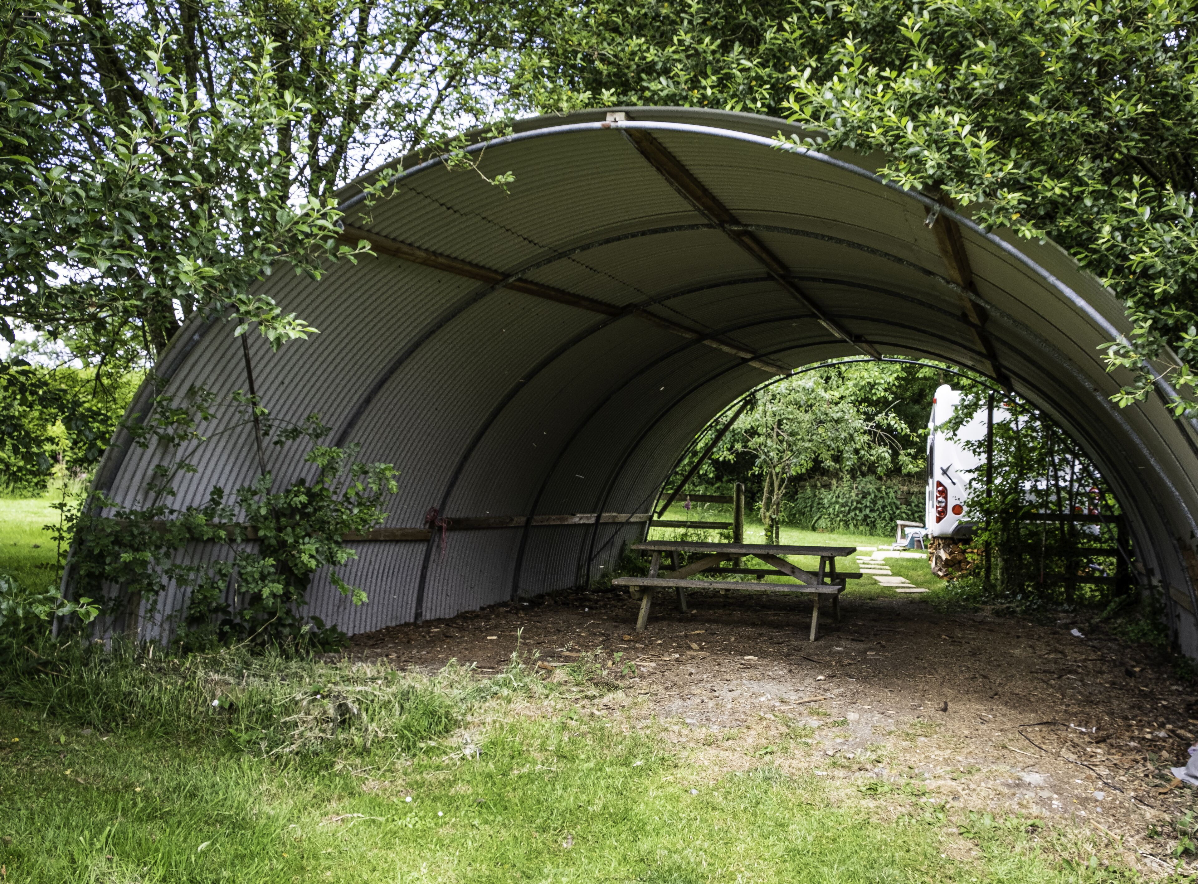 A semi-circular metal shelter with a picnic table in the center, surrounded by grass and trees.
