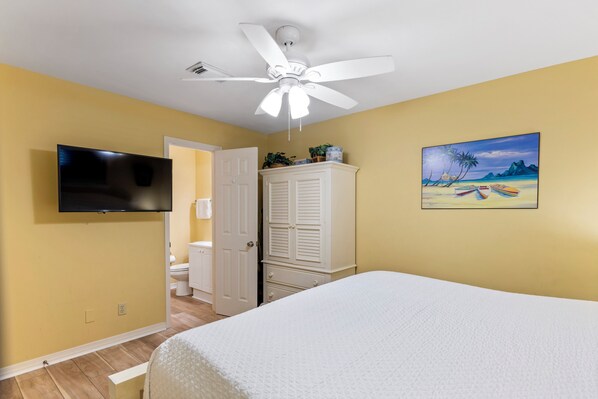 A bedroom with a bed, a ceiling fan, a TV mounted on the wall, and a painting of a beach scene.