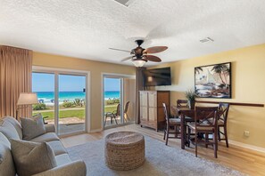 A living room with a view of the ocean, a ceiling fan, a dining area with chairs, and a TV mounted on the wall.
