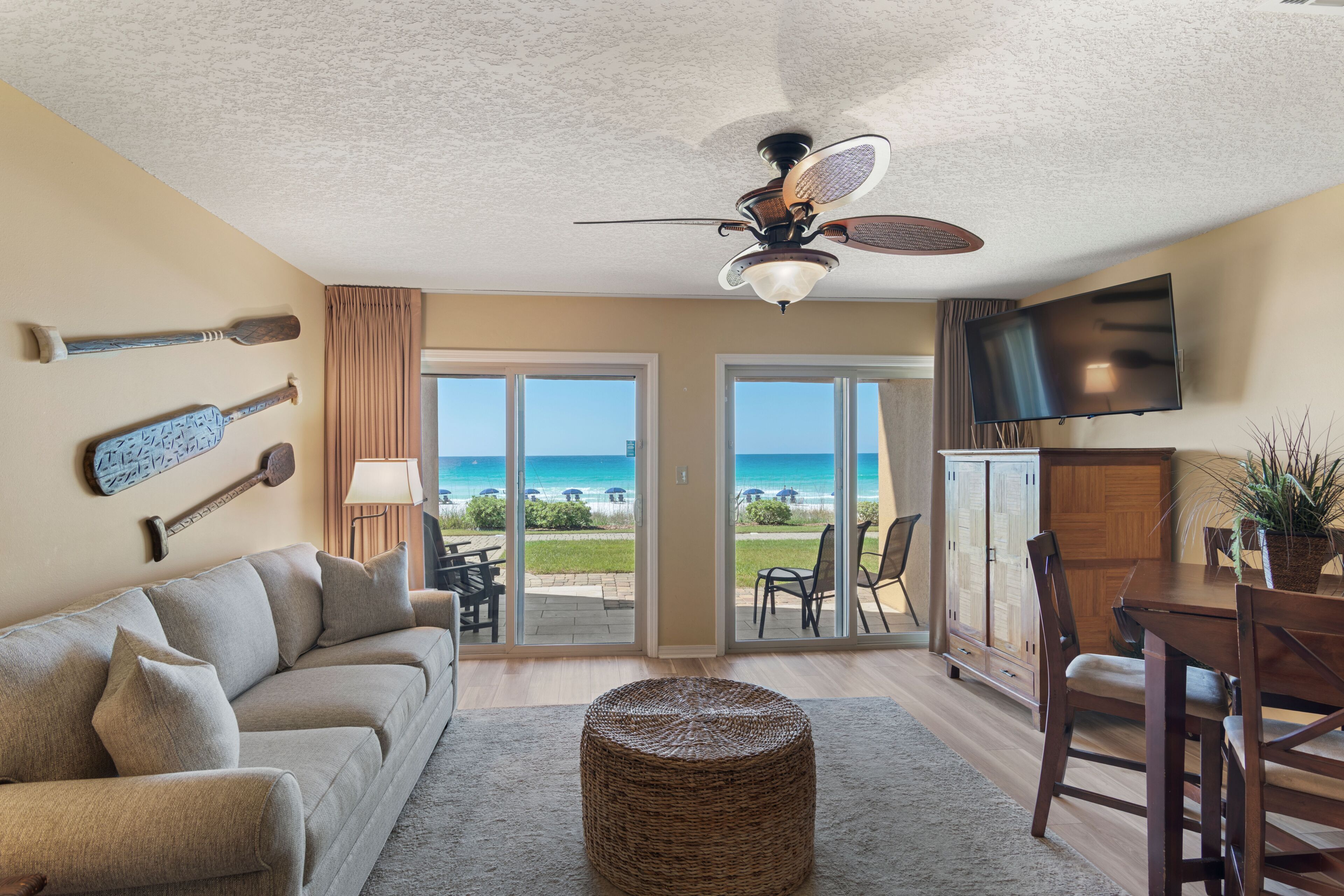 A living room with a sofa, a dining table, chairs, a ceiling fan, a TV, and a view of the ocean through large glass doors.