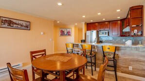 A dining area with a round table and chairs, a kitchen with wooden cabinets and a marble countertop, and artwork on the walls. - Ocean Front Condo Shorebirds 83 (Pajaro Dunes)