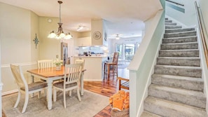 A dining area with a wooden table and chairs, a kitchen in the background, and a staircase with carpeted steps.