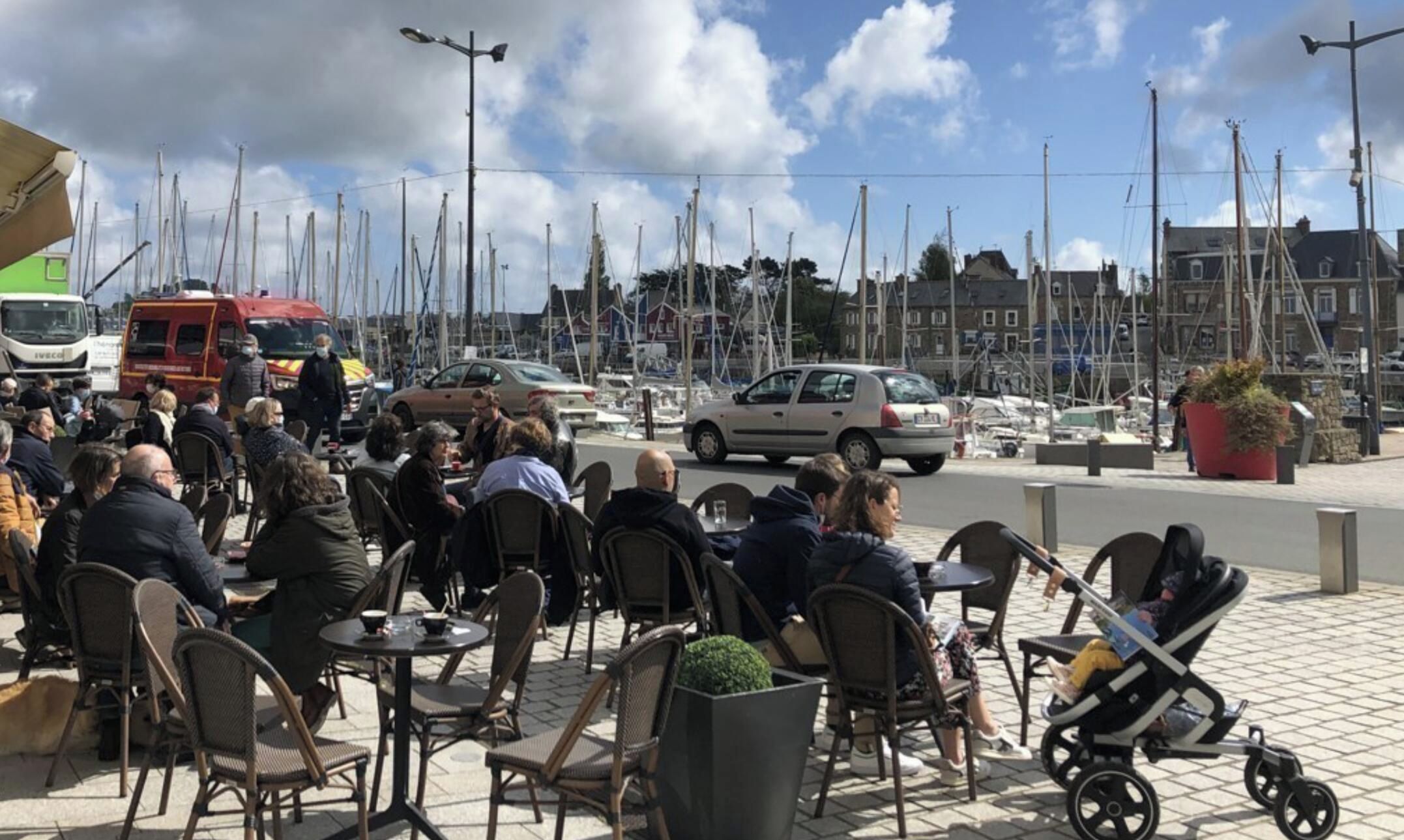 Un espace extérieur aménagé avec des tables et des chaises, une poussette, et une vue sur le port de plaisance, avec des bateaux et des bâtiments en arrière-plan.