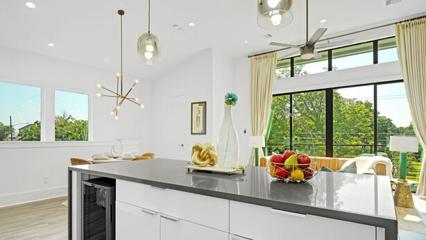 A modern kitchen with a central island, pendant lights, and a view of greenery outside.