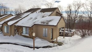 Une maison de plain-pied, avec un toit en pente recouvert de neige, entourée d’une clôture en bois et d’arbres sans feuilles.
