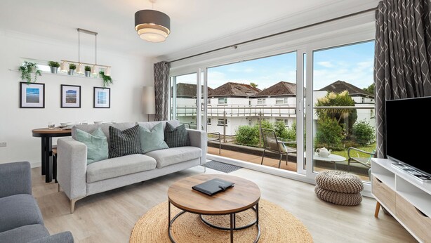 Una sala de estar moderna con un sofá gris, una mesa redonda y un televisor sobre un mueble blanco. Amplias ventanas ofrecen vistas del barrio.