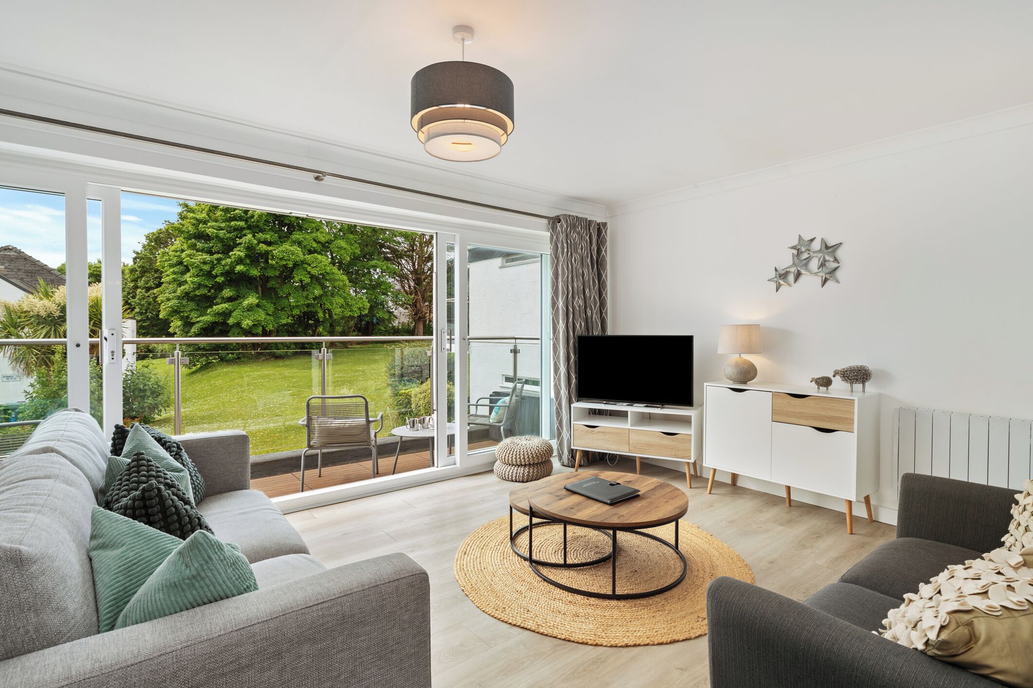 A modern living room with a sofa, coffee table, TV, and a view of a garden through large sliding doors.