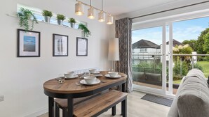 Un comedor moderno con una mesa de madera preparada para cuatro personas, un banco y vistas a un balcón con plantas y una casa al fondo.