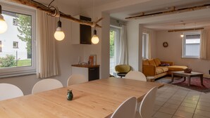 Un saló modern amb una taula de dinar de fusta, cadires blanques, un sofà groc i una taula auxiliar. Hi ha finestres grans amb cortines, una làmpada penjant i una porta ben visible.
