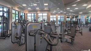 Un gimnasio amplio con diversas máquinas de ejercicio, grandes ventanales y piso alfombrado.