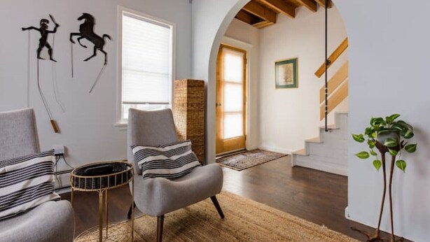 Un salon moderne avec un fauteuil gris, un canapé à rayures et une petite table ronde. Un escalier en bois mène à une autre pièce, et une plante est visible sur le côté droit.