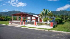 Un edificio con un espacio exterior techado, jardín cercado y una montaña al fondo.