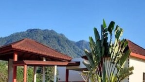 Un edificio de paredes blancas y techo rojo, rodeado de vegetación y montañas al fondo.