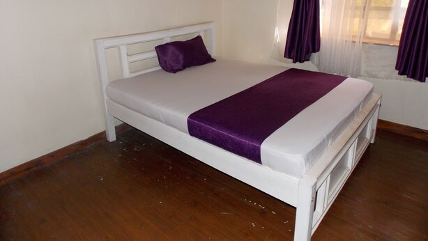 Ein Einzelbett mit weißem Gestell und einer violetten Tagesdecke in einem Zimmer mit Holzboden und einem Fenster mit violetten Vorhängen.
