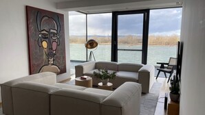 A modern living room with a large window overlooking a body of water, a beige sofa, a floor lamp, and a painting on the wall. - Modernes Apartment mit Wunderschönem Rheinblick (Urmitz)