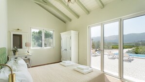 Ein Schlafzimmer mit einem großen Bett, einem Kleiderschrank, einem Schreibtisch mit Lampe sowie Blick auf einen Pool und Berge.