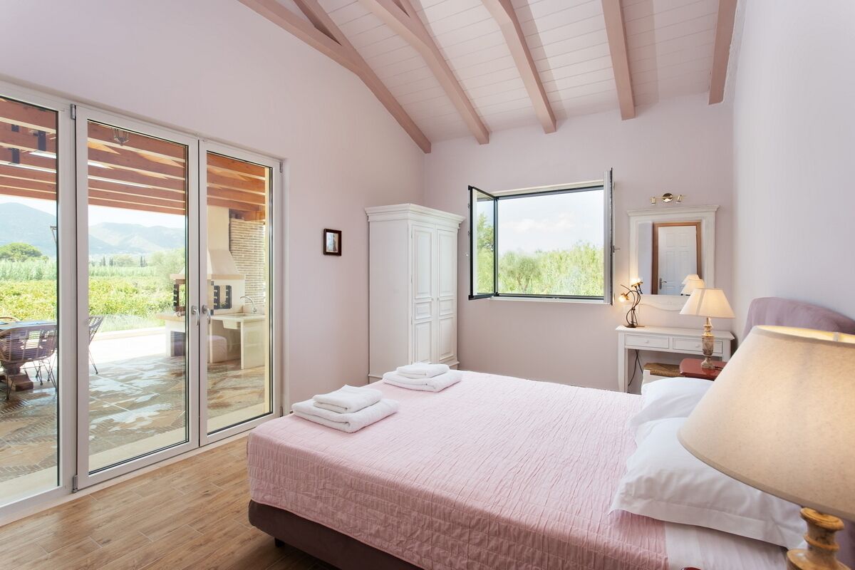 Una camera da letto con un'ampia porta-finestra che dà su una zona esterna, un letto con biancheria rosa, un armadio bianco, un comodino con una lampada e uno specchio appeso al muro.