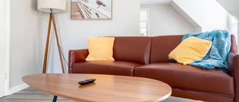 A living room with a brown leather sofa, a round wooden coffee table, a floor lamp, and a framed picture on the wall.