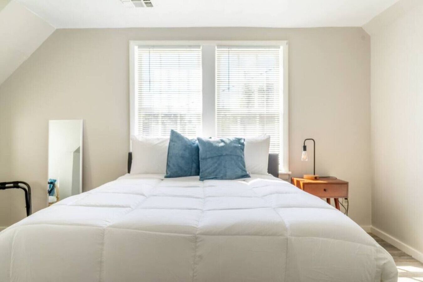 Un dormitorio con una cama grande, dos ventanas con persianas, una mesita de noche con lámpara y un espejo en la pared.
