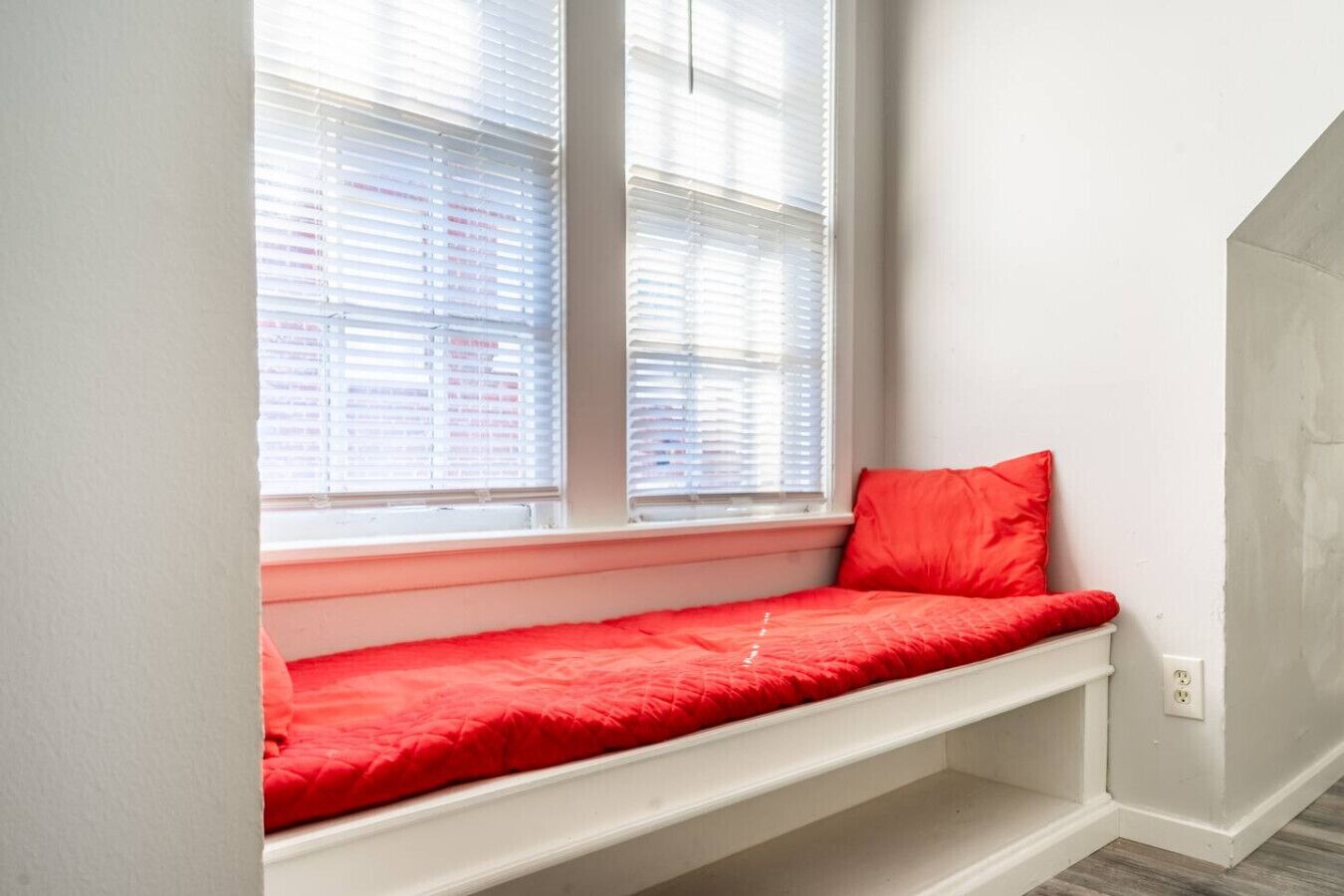 A room with a built-in bench under a window, white walls, and a visible electrical outlet.